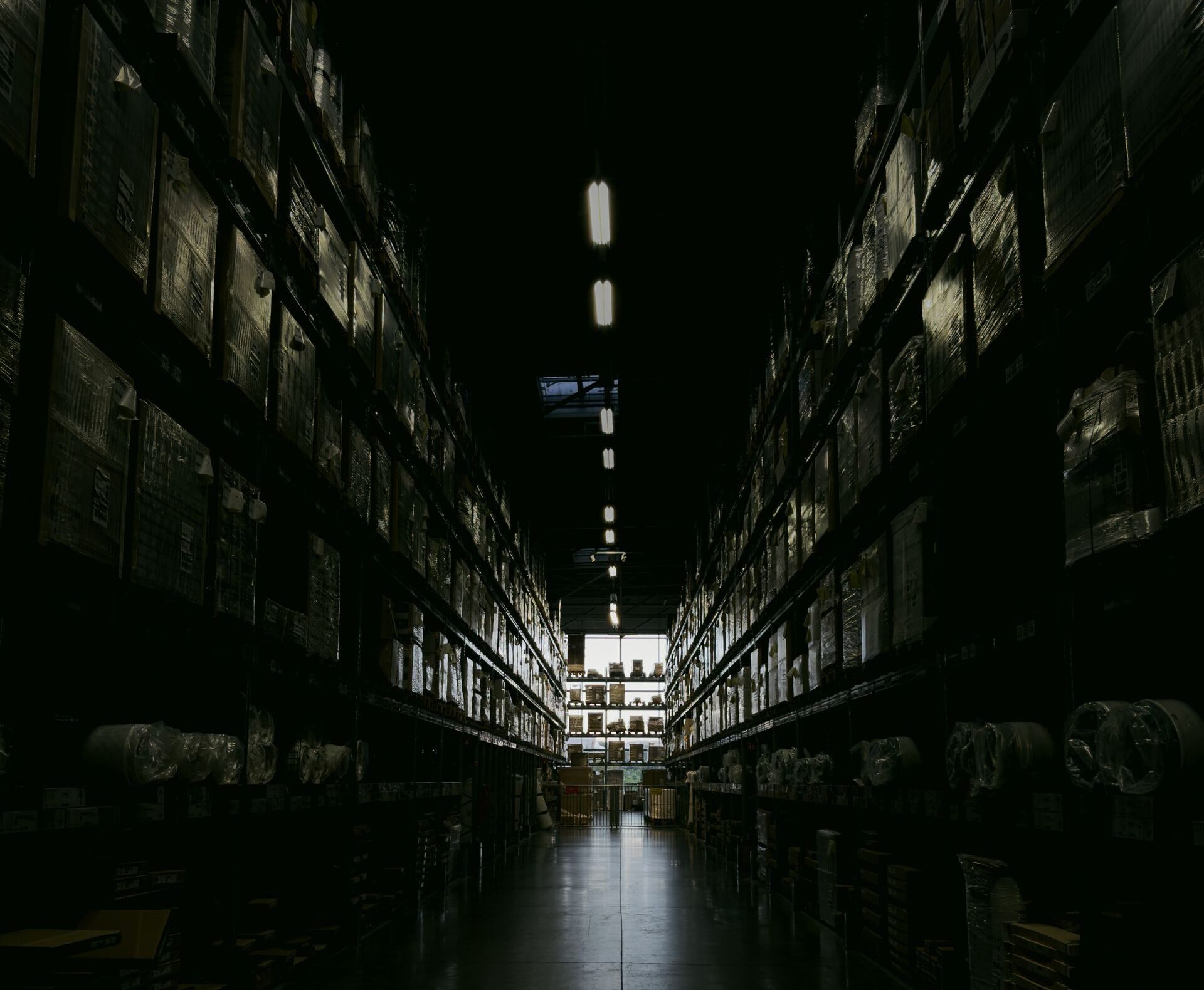 A dark, narrow warehouse aisle with shelves filled with boxes and goods, illuminated by overhead lights.